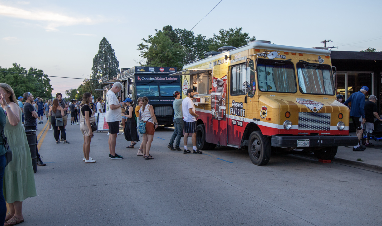 People stand in line at two food trucks parked on a street during the day.